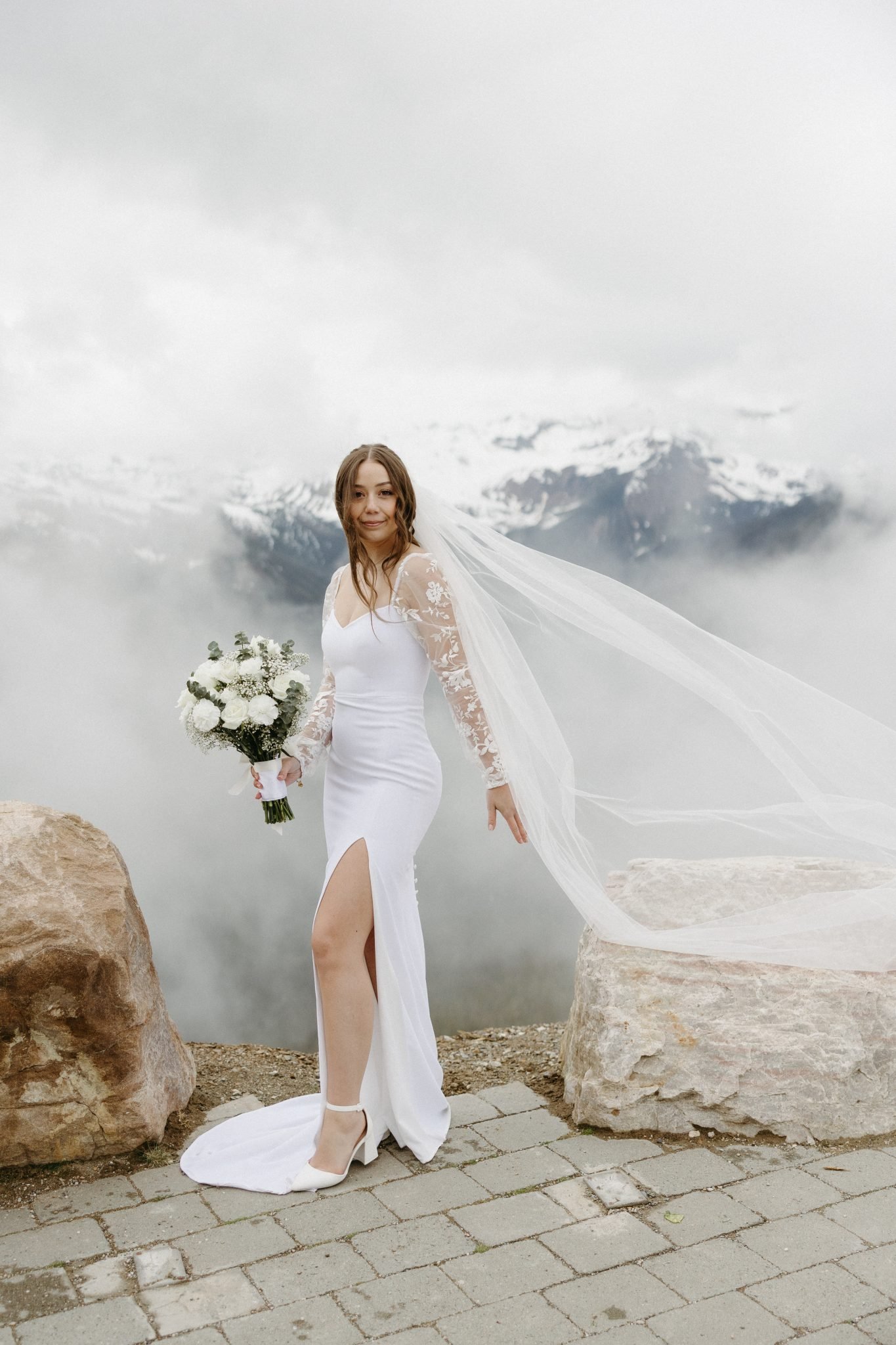 Bride in modern white gown with lace sleeves stands on a mountaintop with flowing veil during a Golden british columbia spring wedding rocky mountain bride.