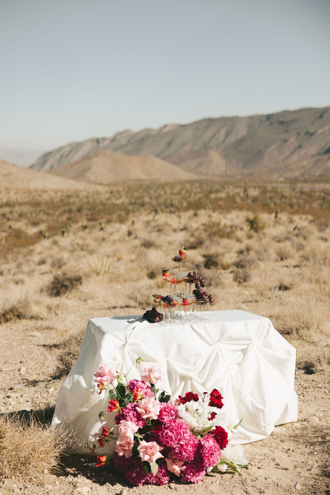Champagne tower styled on a white-draped table with bright pink and red florals set against a desert backdrop for the Riann Hassell styled shoot las vegas nevada rocky mountain bride.