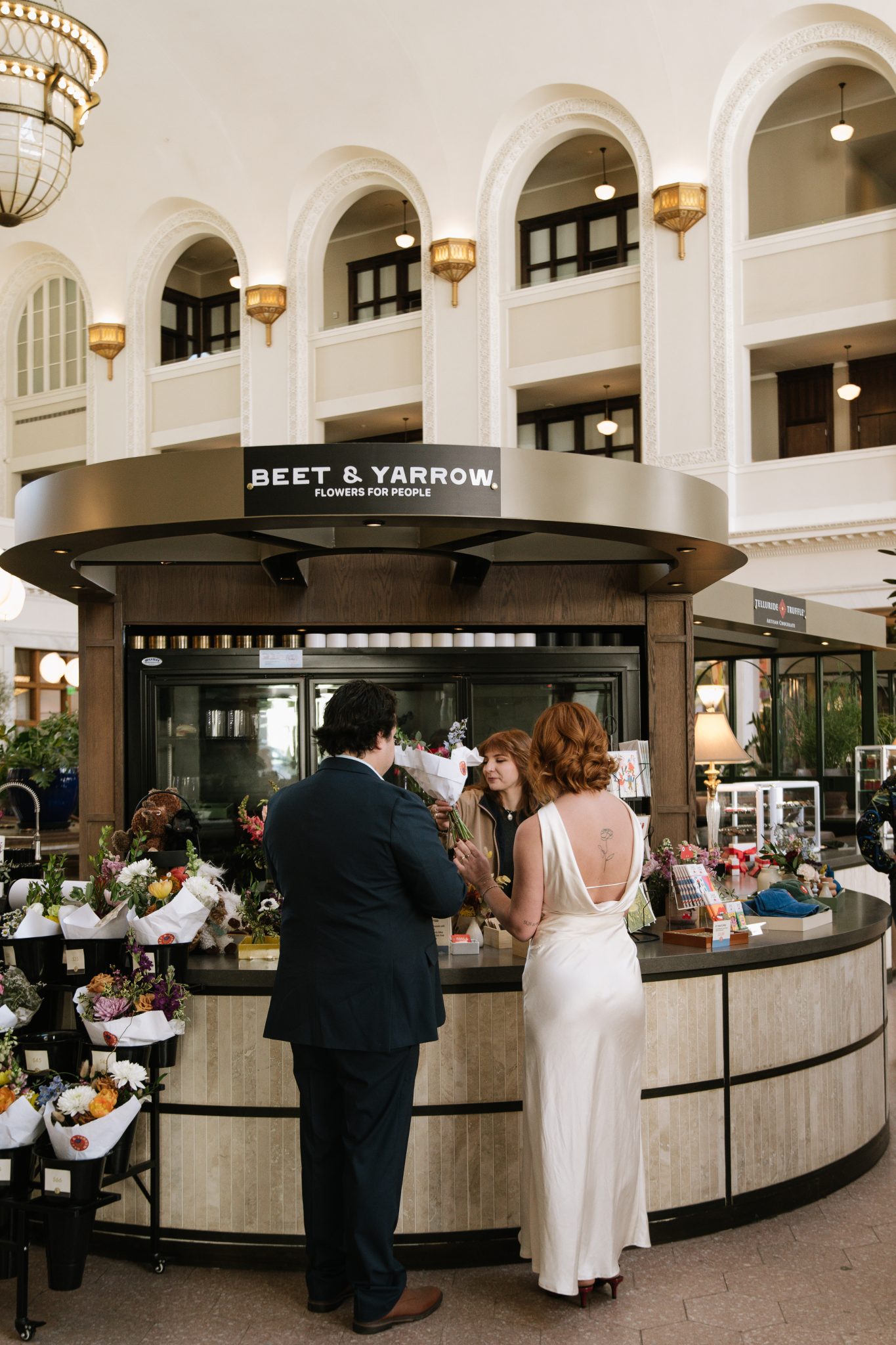 A bride in a satin dress and groom in a navy suit select a bouquet at a flower shop kiosk inside a historic building for their Salt Photography engagement rocky mountain bride shoot.