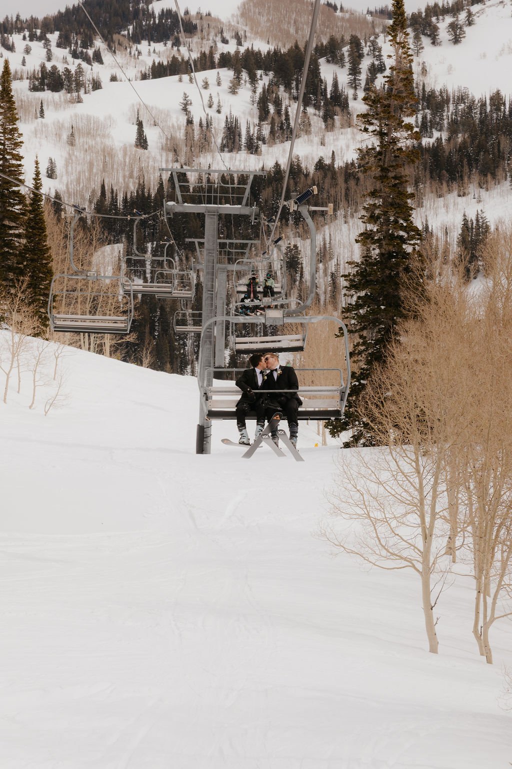Two grooms share a kiss on a ski lift surrounded by snow-covered trees during their Spring Brighton Resort elopement.