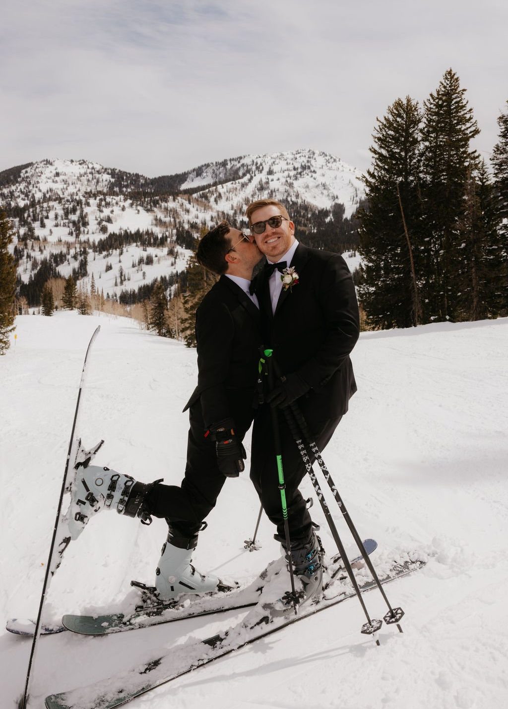 One groom kisses the other on the cheek while standing in skis with snow-covered mountains behind them at their Spring Brighton Resort elopement.