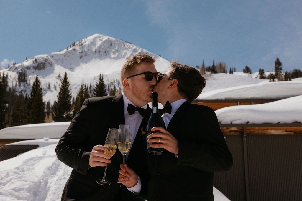 Grooms in black tuxedos kiss while holding champagne and a bottle in front of a snowy mountain backdrop during their Spring Brighton Resort elopement.