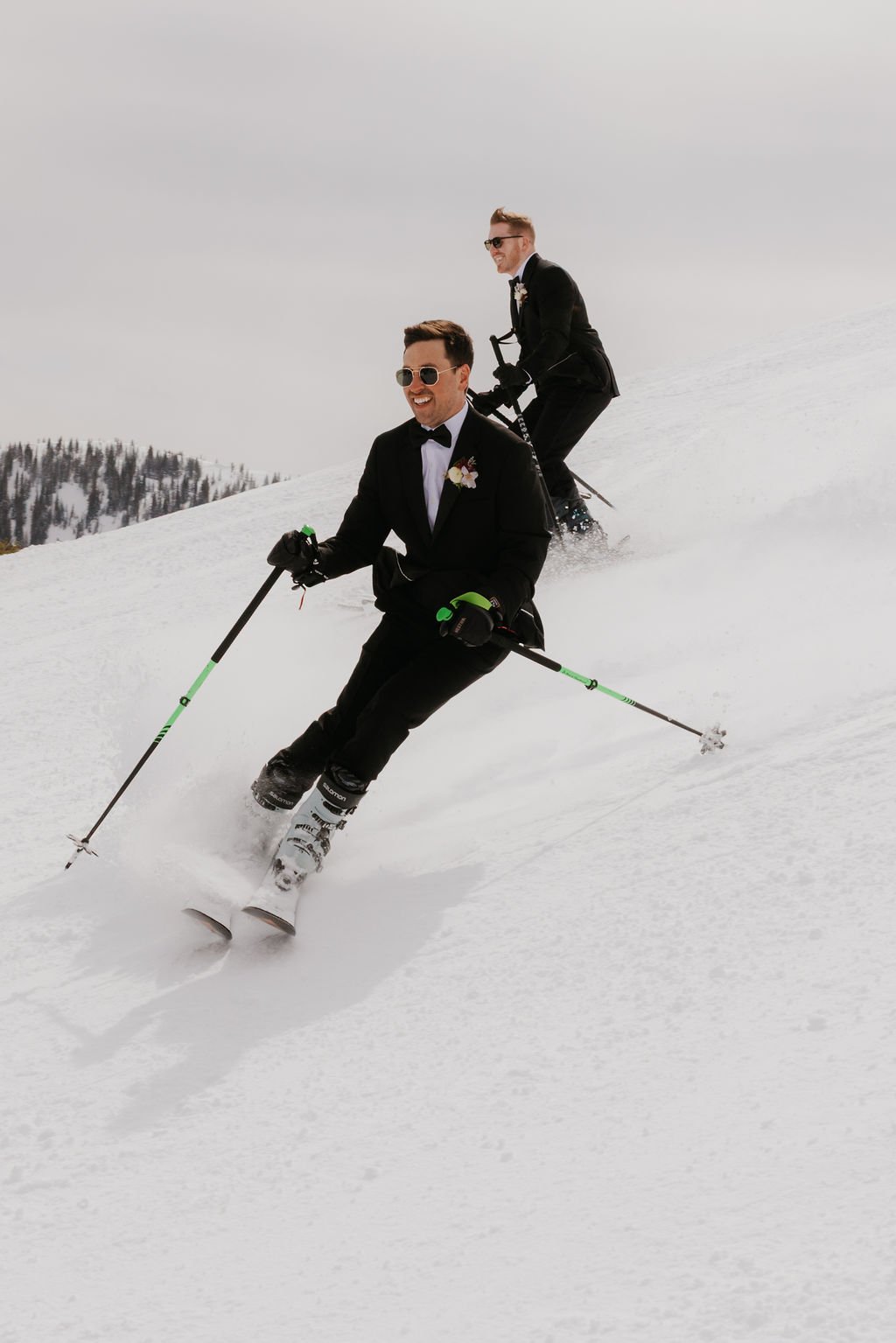 Grooms skiing down a mountain slope in tuxedos on a sunny day for their Spring Brighton Resort elopement.