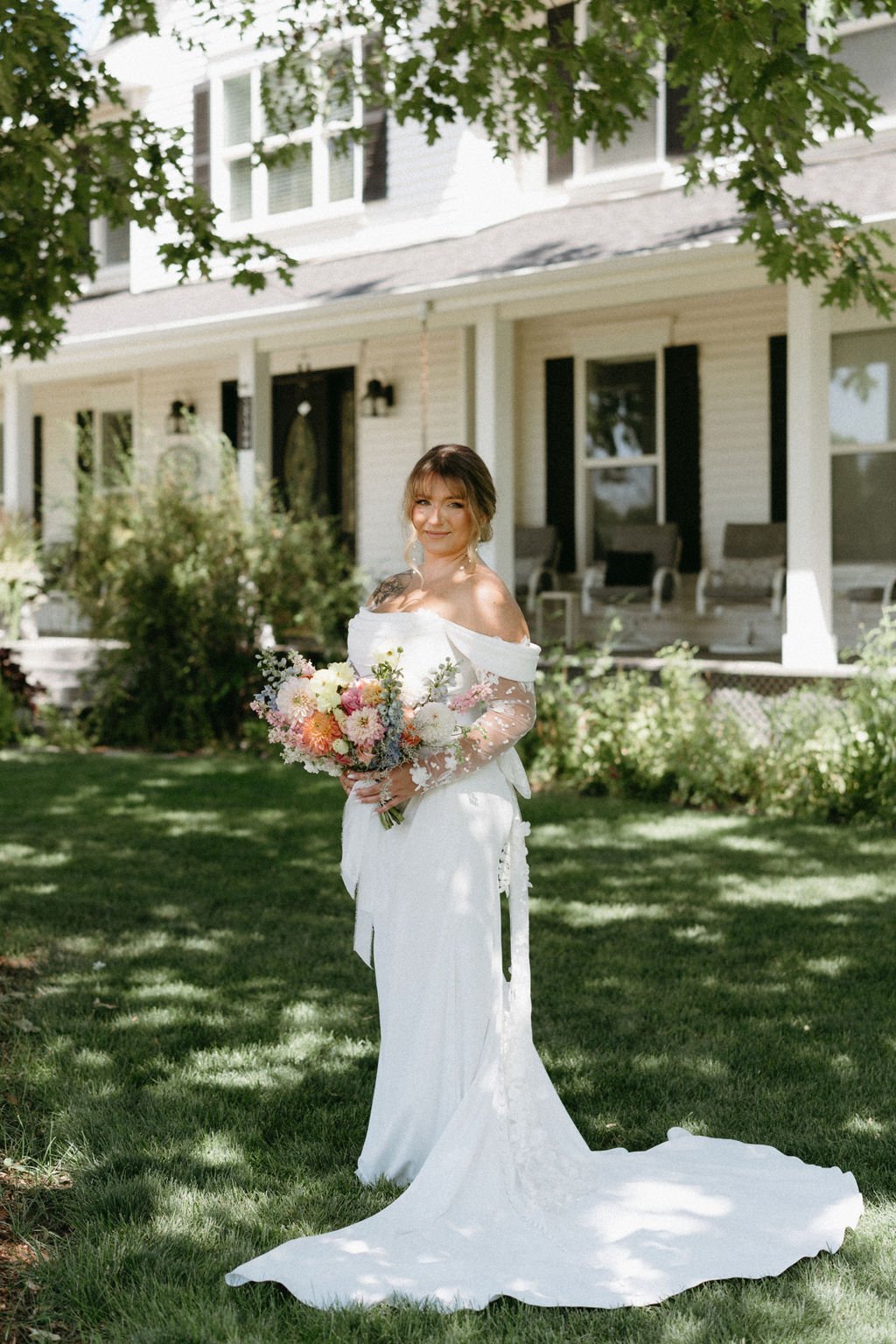 Bride in an off-the-shoulder gown holding a vibrant garden bouquet in front of a farmhouse at Willow Creek Wedding and Events Idaho rocky mountain bride.
