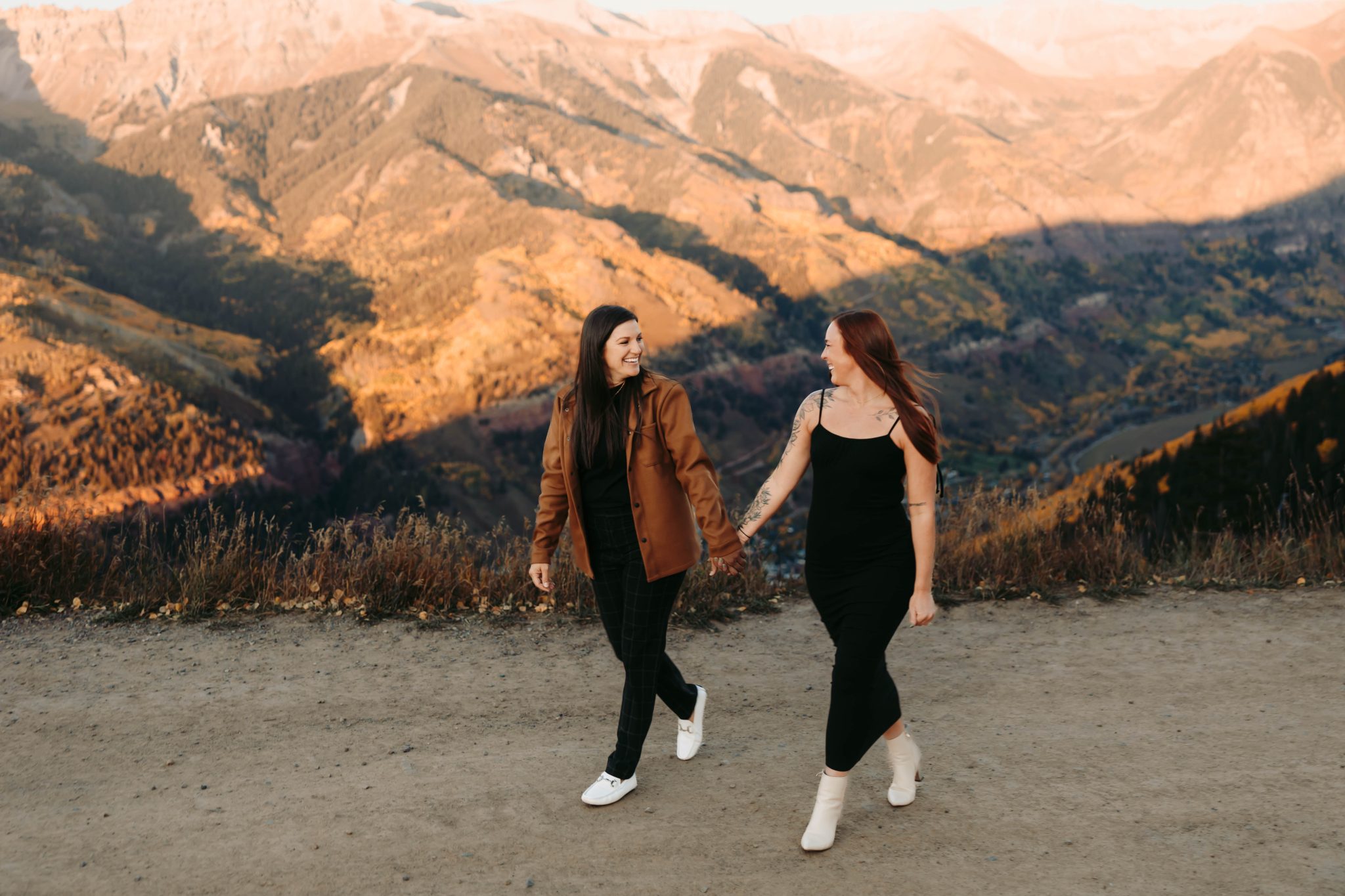 Two women holding hands and walking at the edge of a mountain vista during golden hour for a fall telluride proposal lgbtq+ rocky mountain bride shoot.