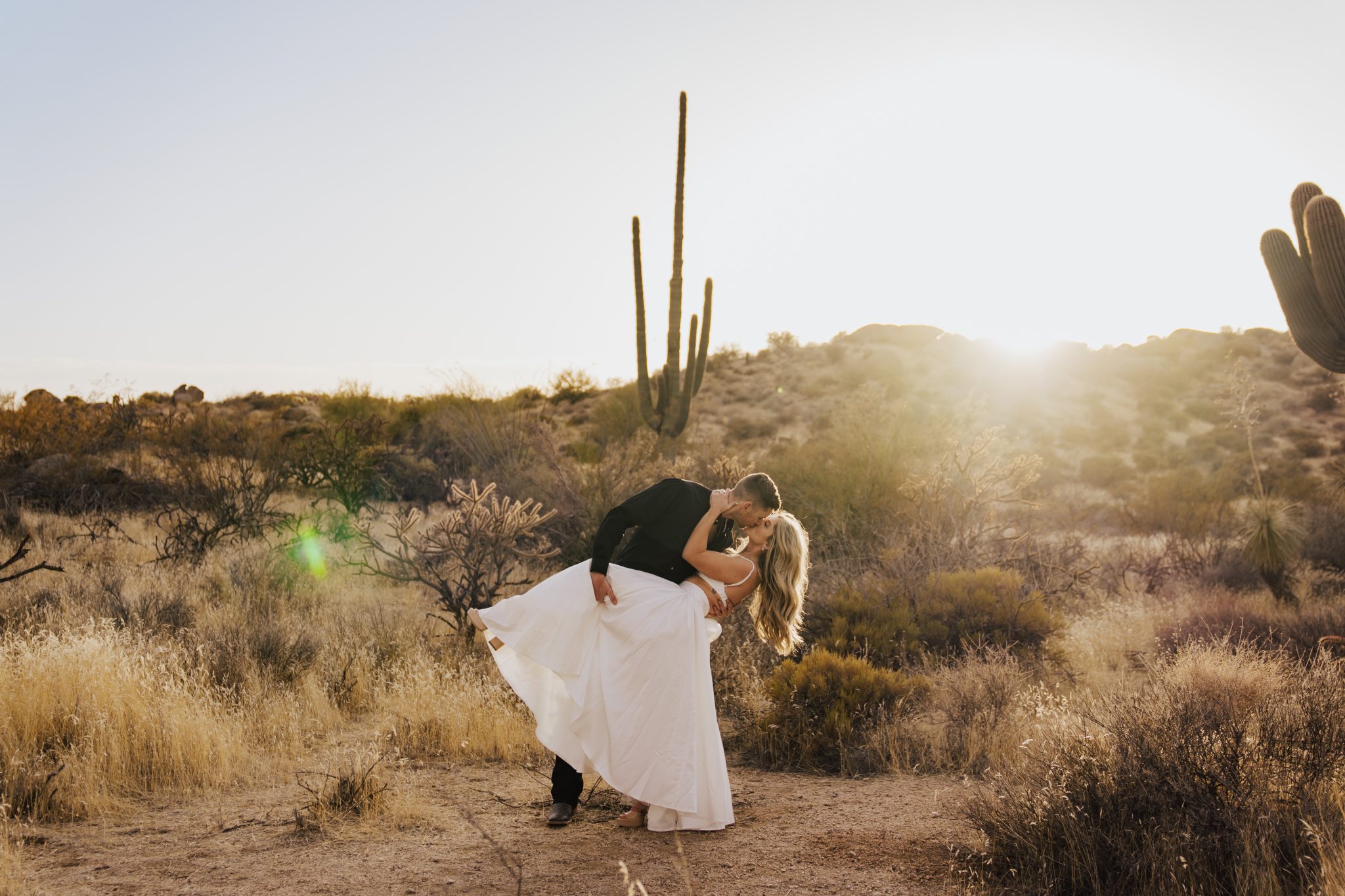 Couple sharing a dip kiss in a flowing white dress at golden hour in their Arizona desert engagement session rocky mountain bride photo shoot.