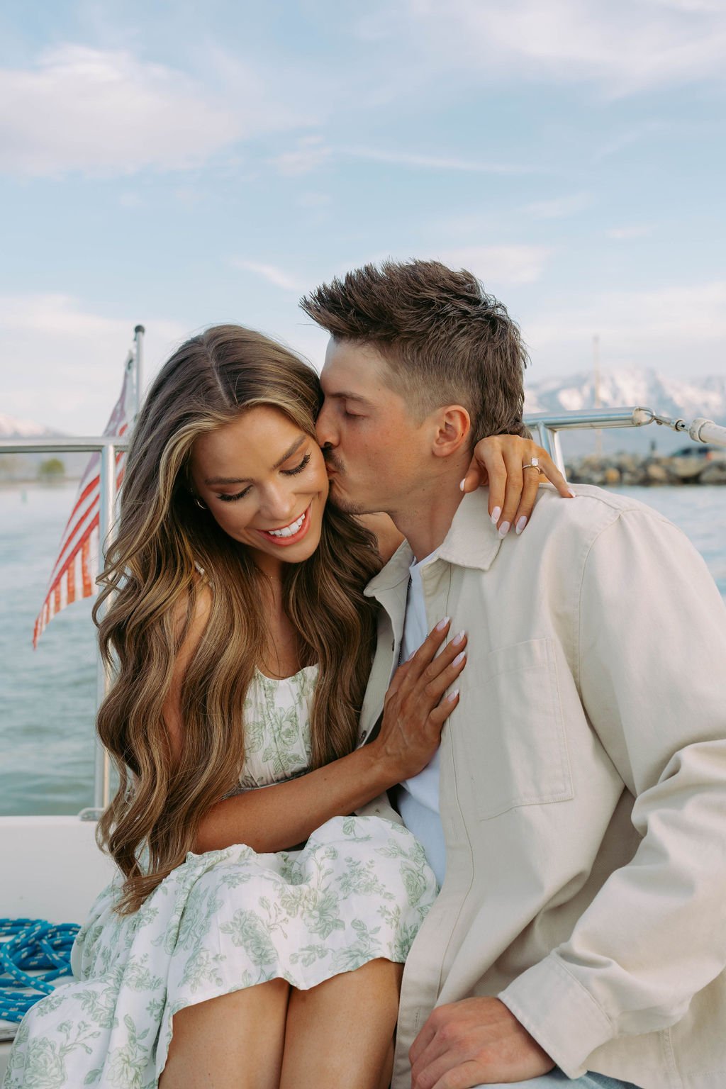A groom-to-be kisses his fiancée on the cheek while she smiles lovingly during a romantic boat ride at sunset, captured in this Bay and Bre Photo Utah engagement session rocky mountain bride.