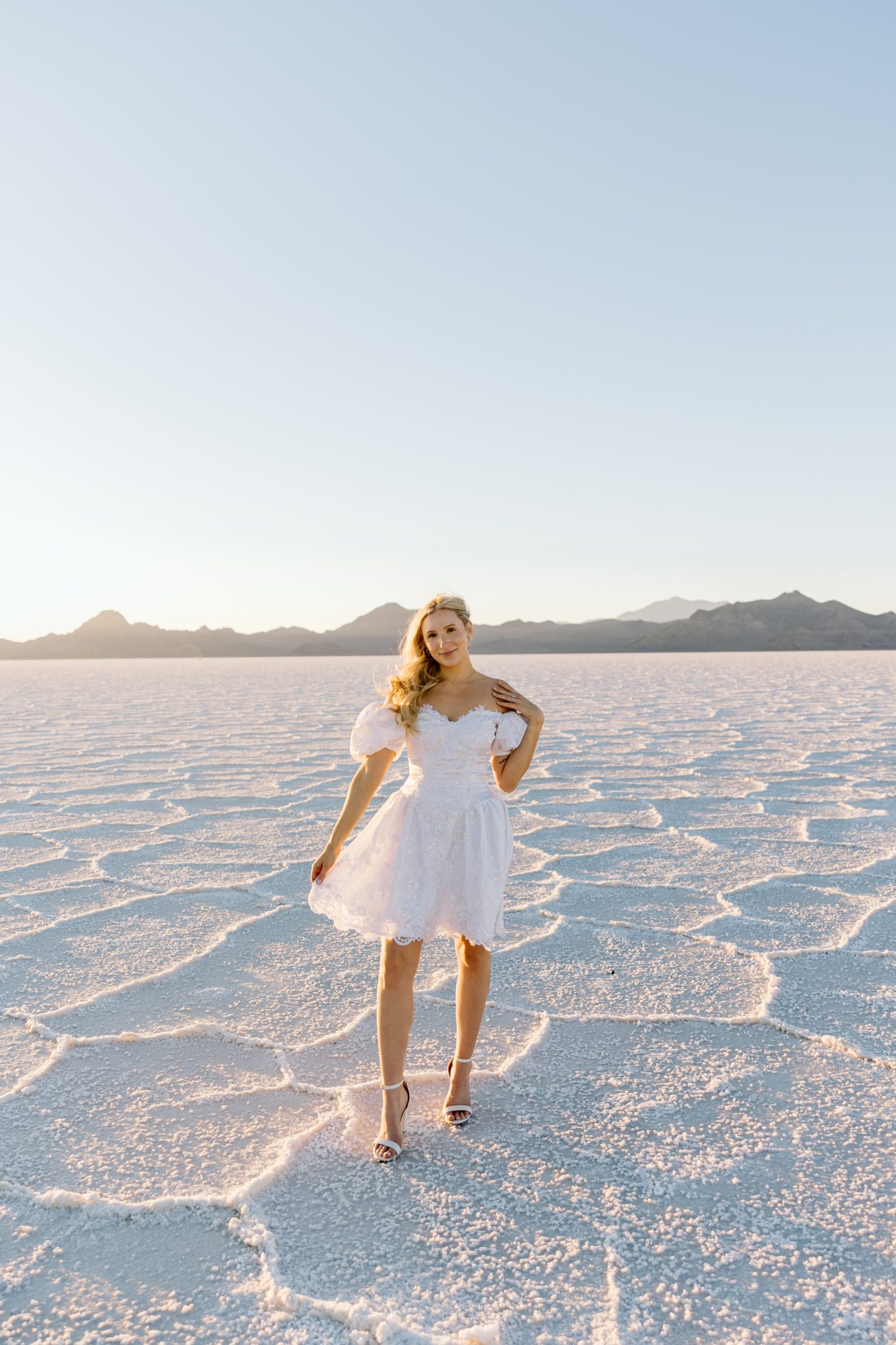 Bride in a white puff-sleeve dress standing alone at sunset during a Bonneville Salt Flats engagement session Niki Baldwin Photography rocky mountain bride.
