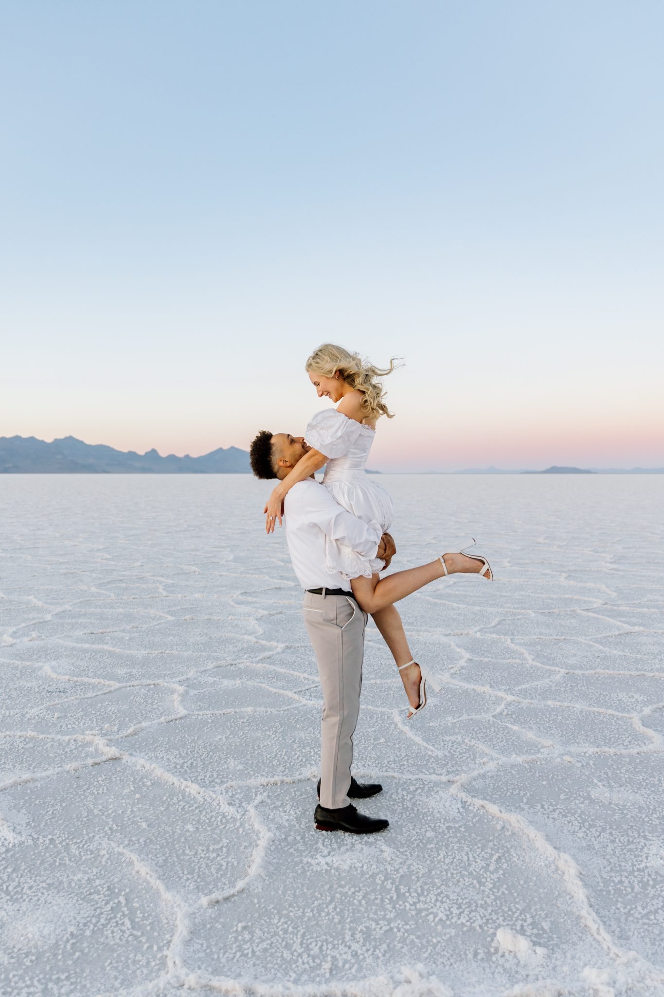 Groom lifting bride in a joyful moment with mountain views during a Bonneville Salt Flats engagement session Niki Baldwin Photography rocky mountain bride.