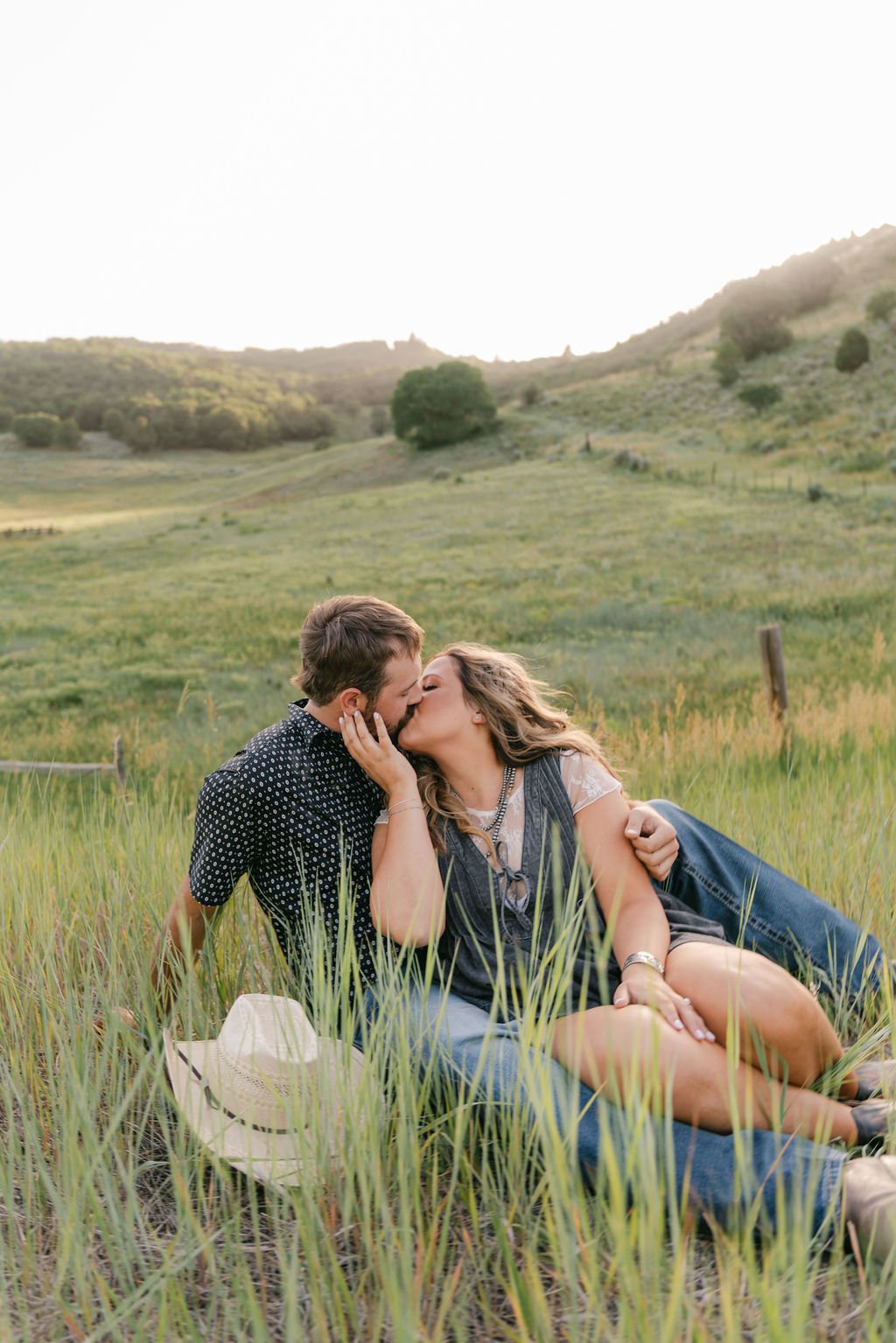 A couple shares a kiss while sitting in tall summer grass during golden hour, captured by Christensen Edits Photography Utah engagement session rocky mountain bride.
