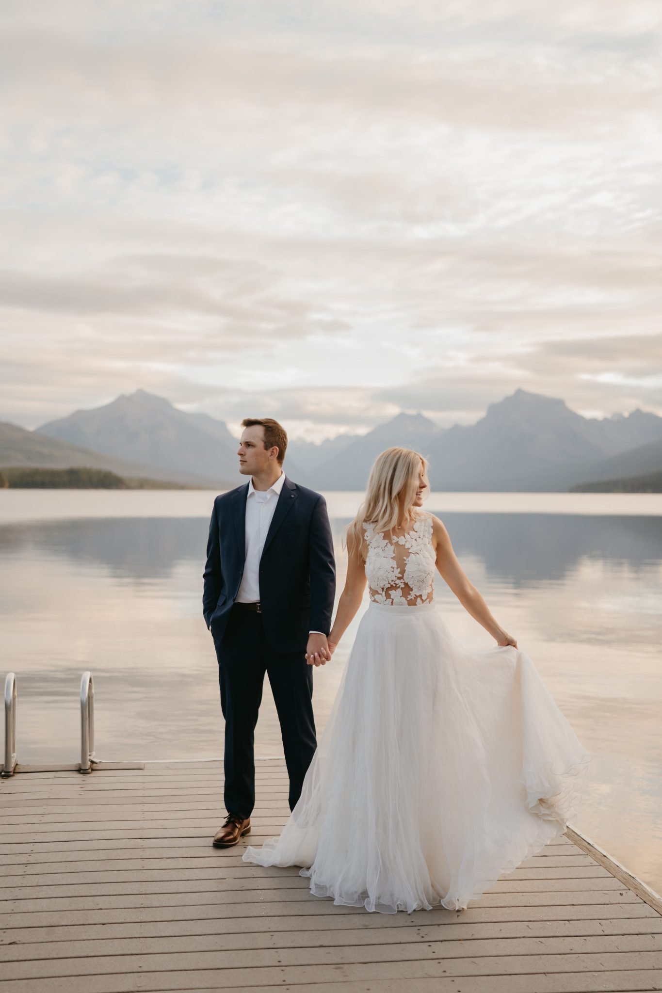 Bride and groom hold hands on a peaceful dock surrounded by majestic peaks in this Kylie Farmer Photography Glacier National Park elopement rocky mountain bride moment.