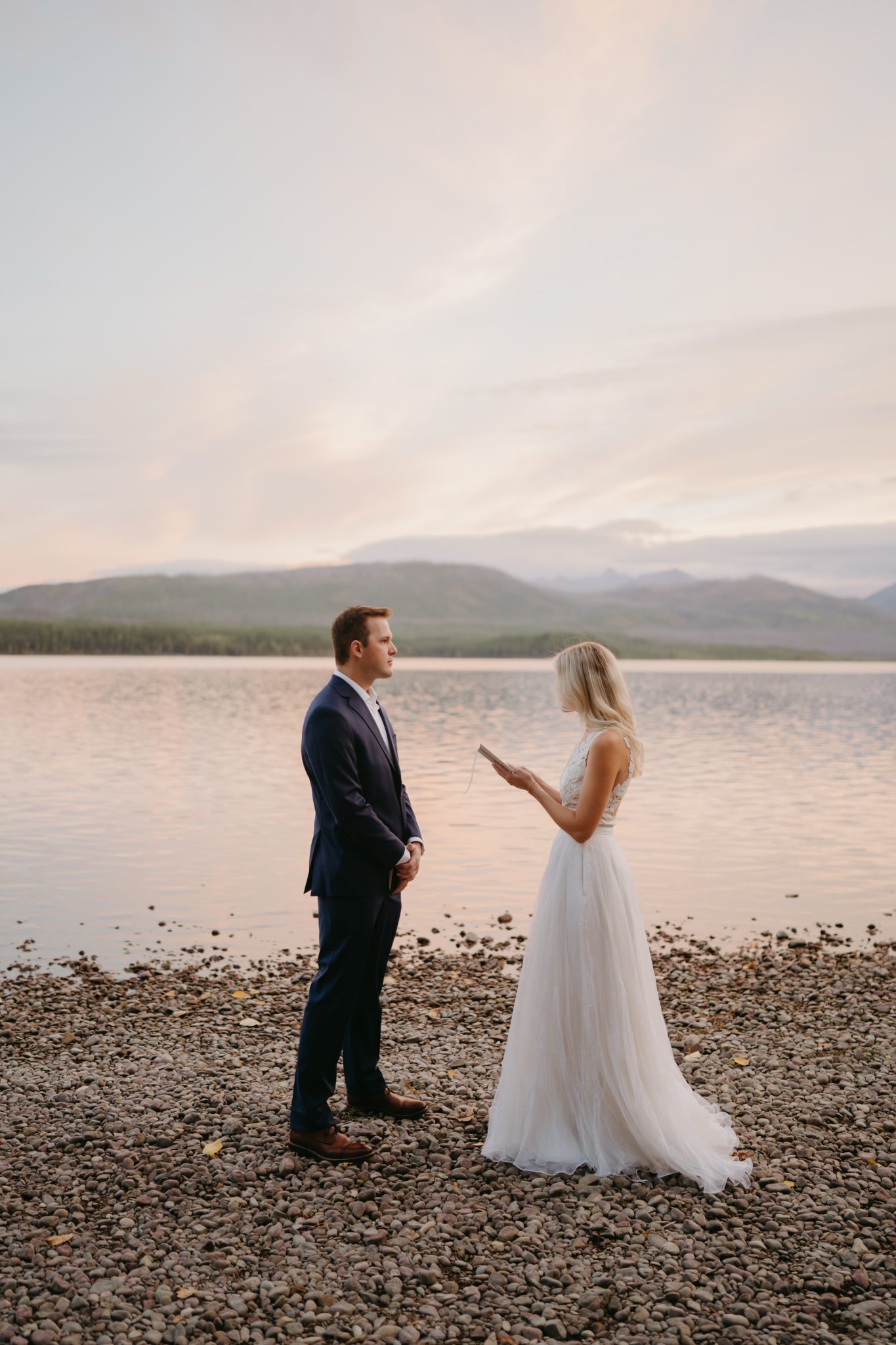 Bride reads vows to groom on a serene lakeshore at sunset captured by Kylie Farmer Photography Glacier National Park elopement rocky mountain bride.