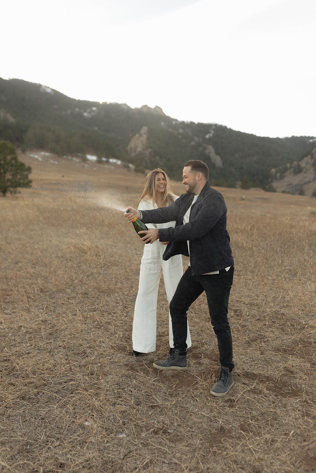 A couple celebrates with champagne in an open mountain field during their S Lynne Photography Boulder Colorado engagement session rocky mountain bride.