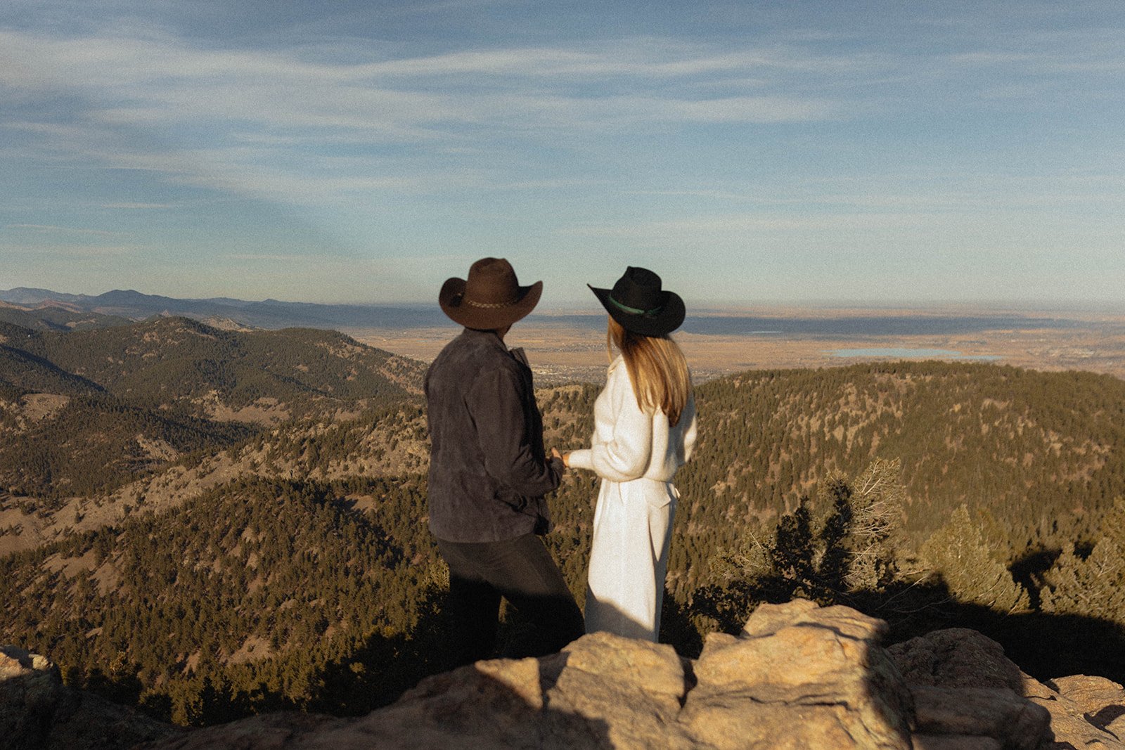 A couple stands hand-in-hand on a mountaintop overlooking a vast valley, captured during their S Lynne Photography Boulder Colorado engagement session rocky mountain bride.