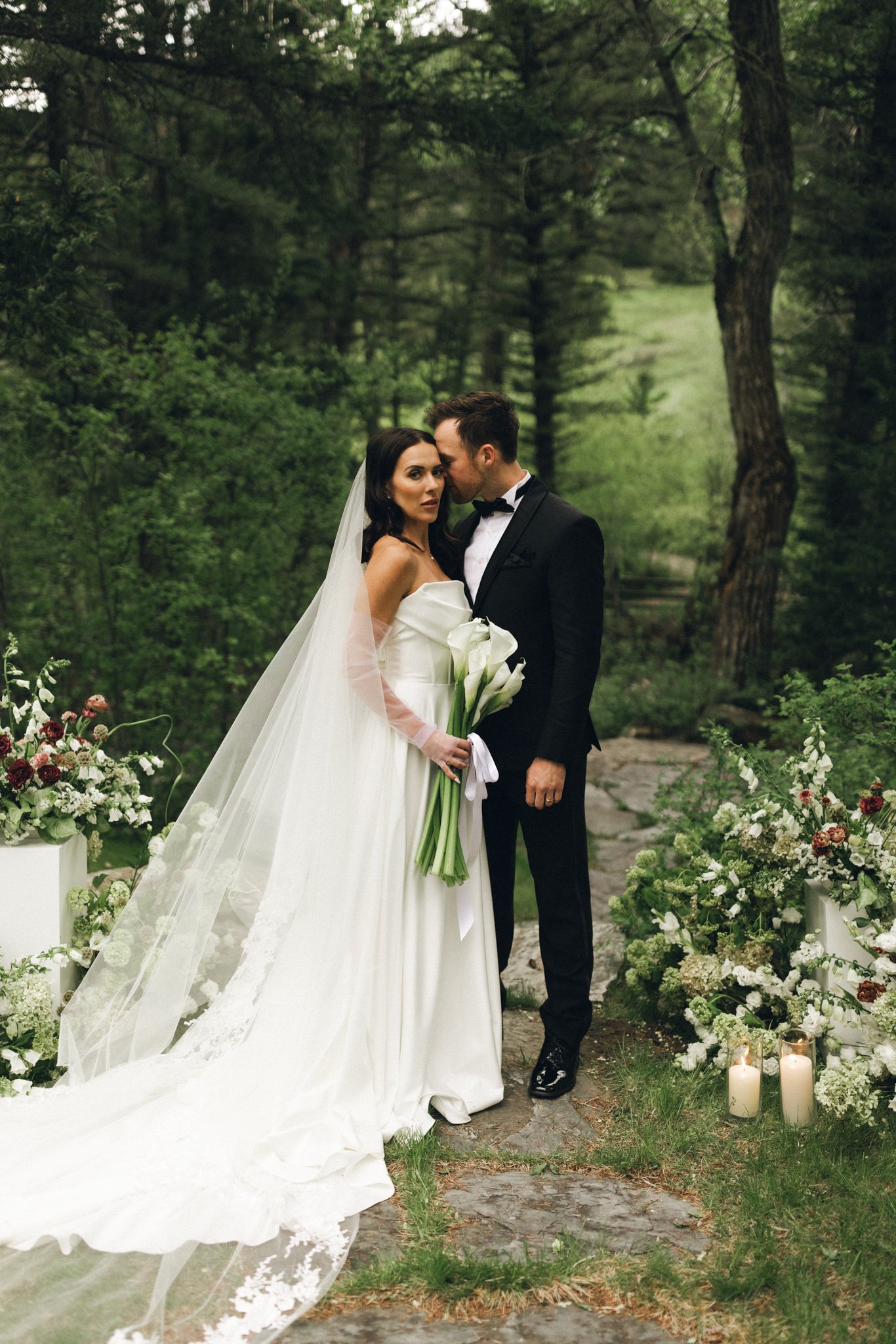 Bride in a structured off-the-shoulder gown and groom in a black tux standing in a forest ceremony space at a Sanctuary at Crow Hollow Ranch wedding inspiration rocky mountain bride shoot.