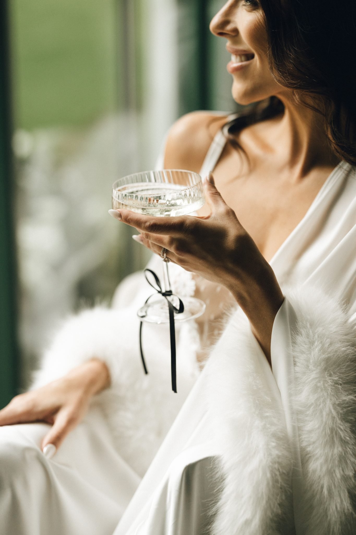 Bride in a white robe with feather trim holding a vintage coupe glass with a black ribbon at a Sanctuary at Crow Hollow Ranch wedding inspiration rocky mountain bride morning moment.
