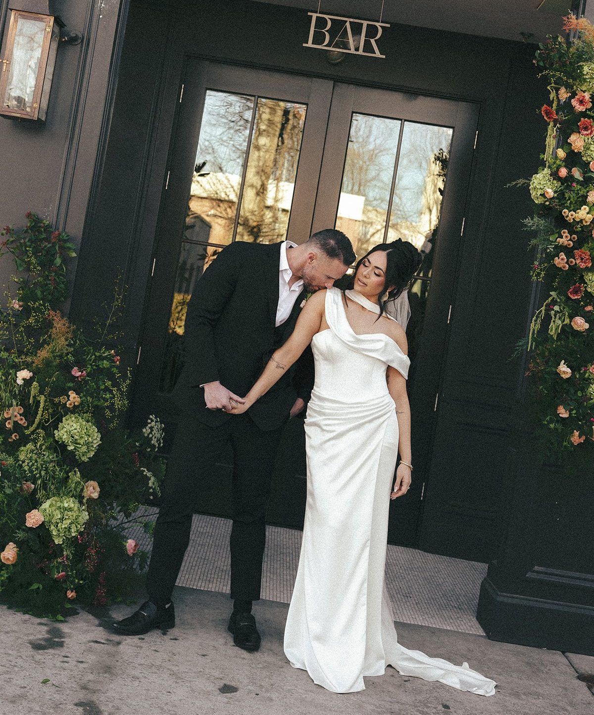 Stylish bride in satin gown and groom in black suit walking past The Lively’s floral-covered exterior for an editorial bride modern tablescape chic wedding the lively downtown boise idaho floral heavy intimate reception dinner pink film elopement.