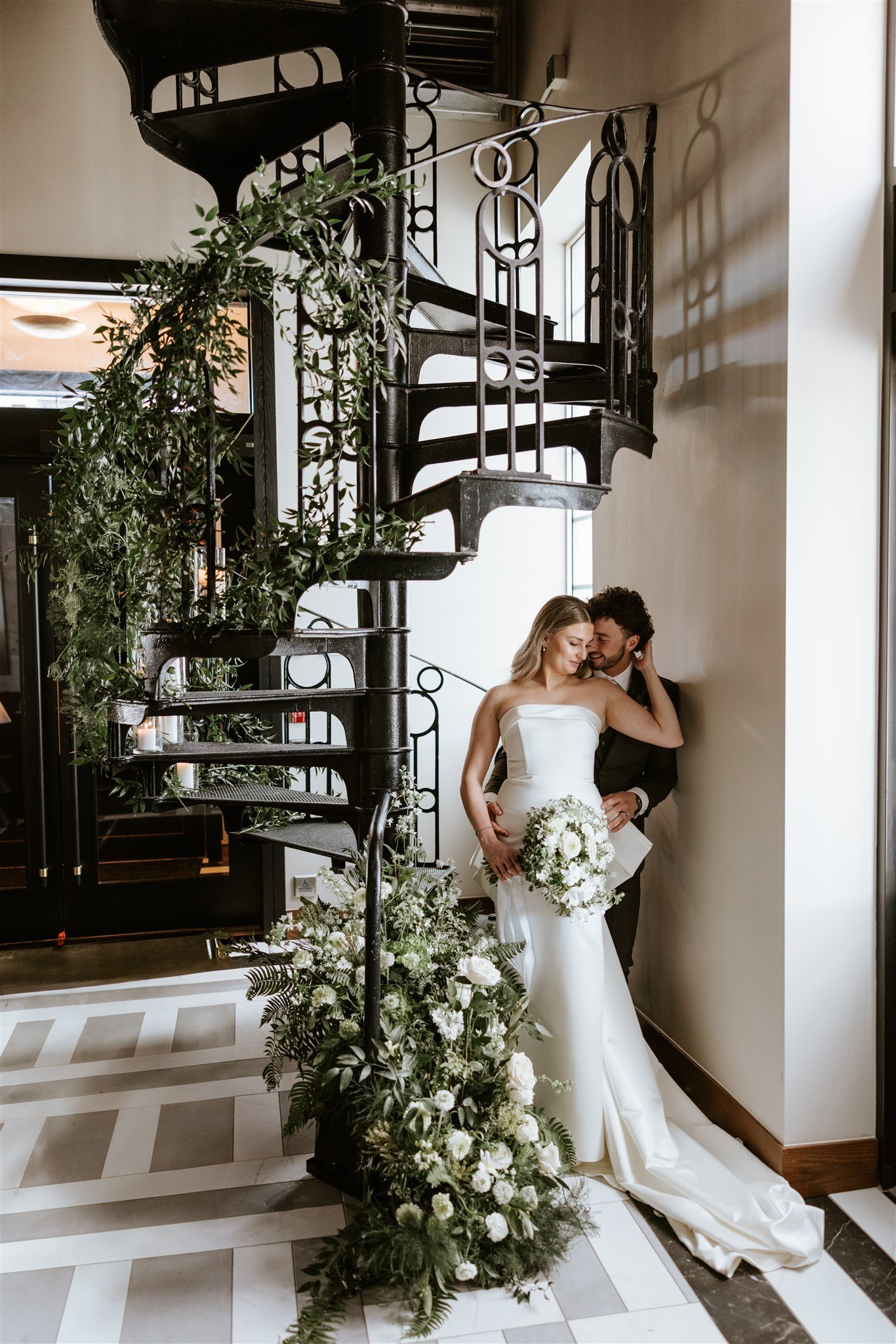 Bride and groom embrace beside romantic spiral staircase with lush greenery during True Wild Distilling styled shoot rocky mountain bride