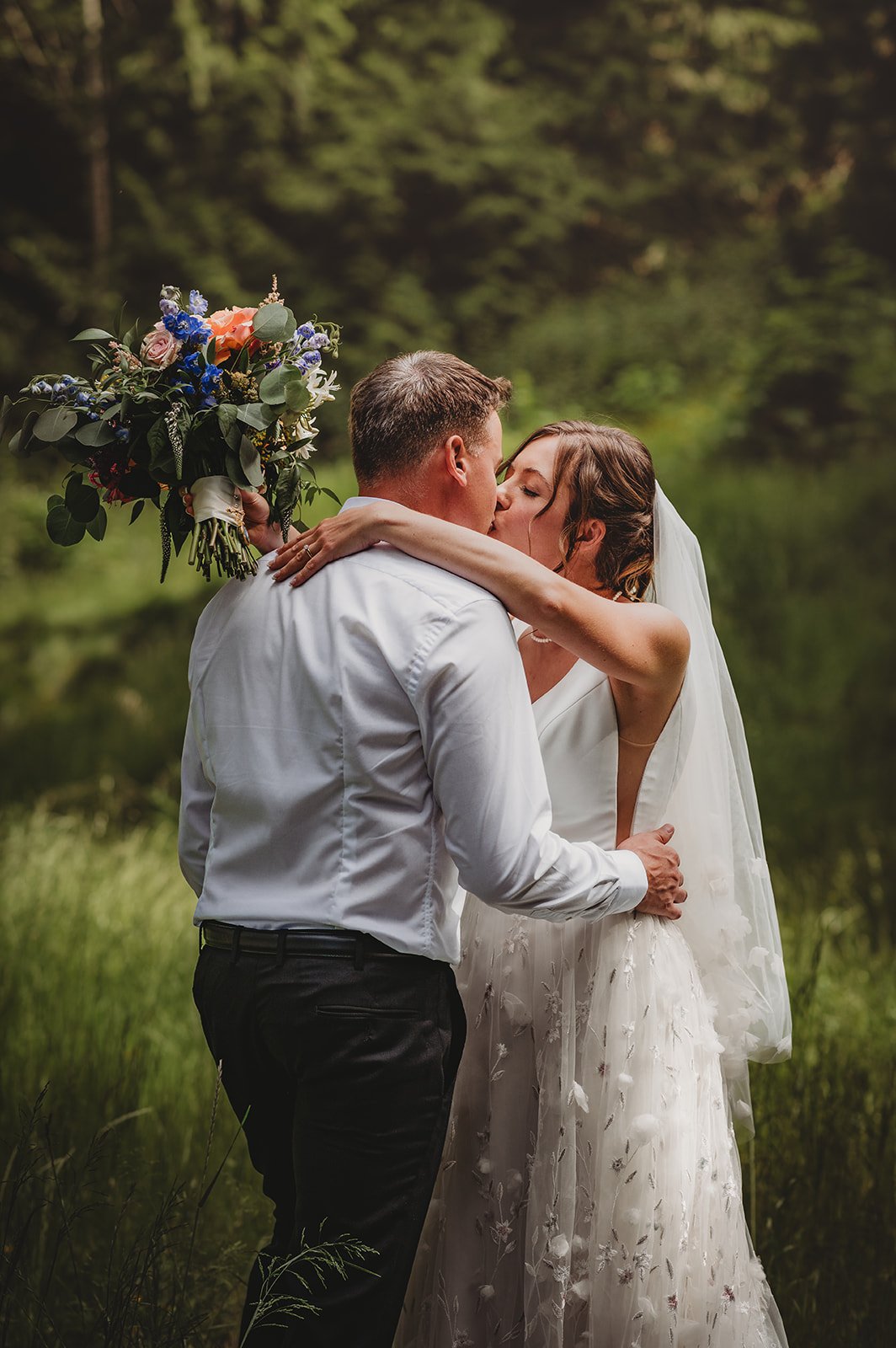 Bride and groom share a romantic kiss in a forest setting holding a vibrant bouquet at a private estate wedding in British Columbia Rocky Mountain Bride.