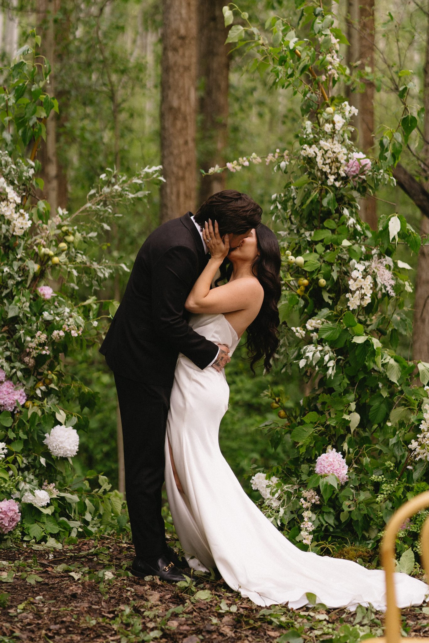 Bride and groom sharing a kiss in a lush forest ceremony surrounded by floral arches — Aurora Valley Farm wedding inspiration british columbia rocky mountain bride