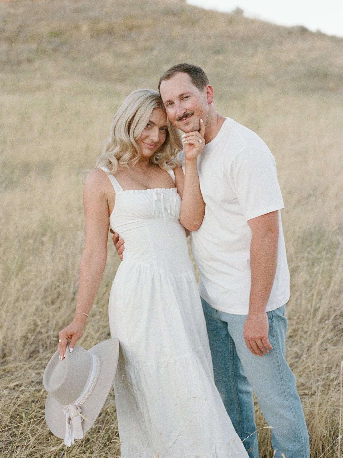 Couple in Idaho's fields wearing matching attire as she hold her neutral color hat in one hand and hold his face in another hand for a Boise Idaho engagement session with Kayla Lucio Photo rocky mountain bride