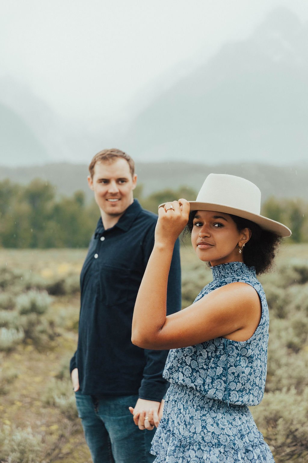 Fiancée adjusting hat while partner smiles behind her at Grand Teton National Park engagement session with Ashlyn French Photography rocky mountain bride