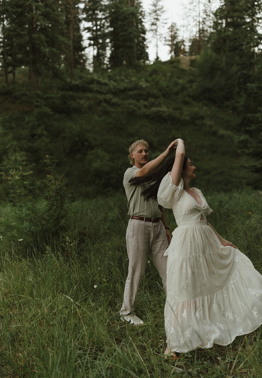 Groom twirling bride in a flowing white dress during a playful moment in the forest clearing, taken for Hanomaly Photo Idaho engagement session rocky mountain bride.