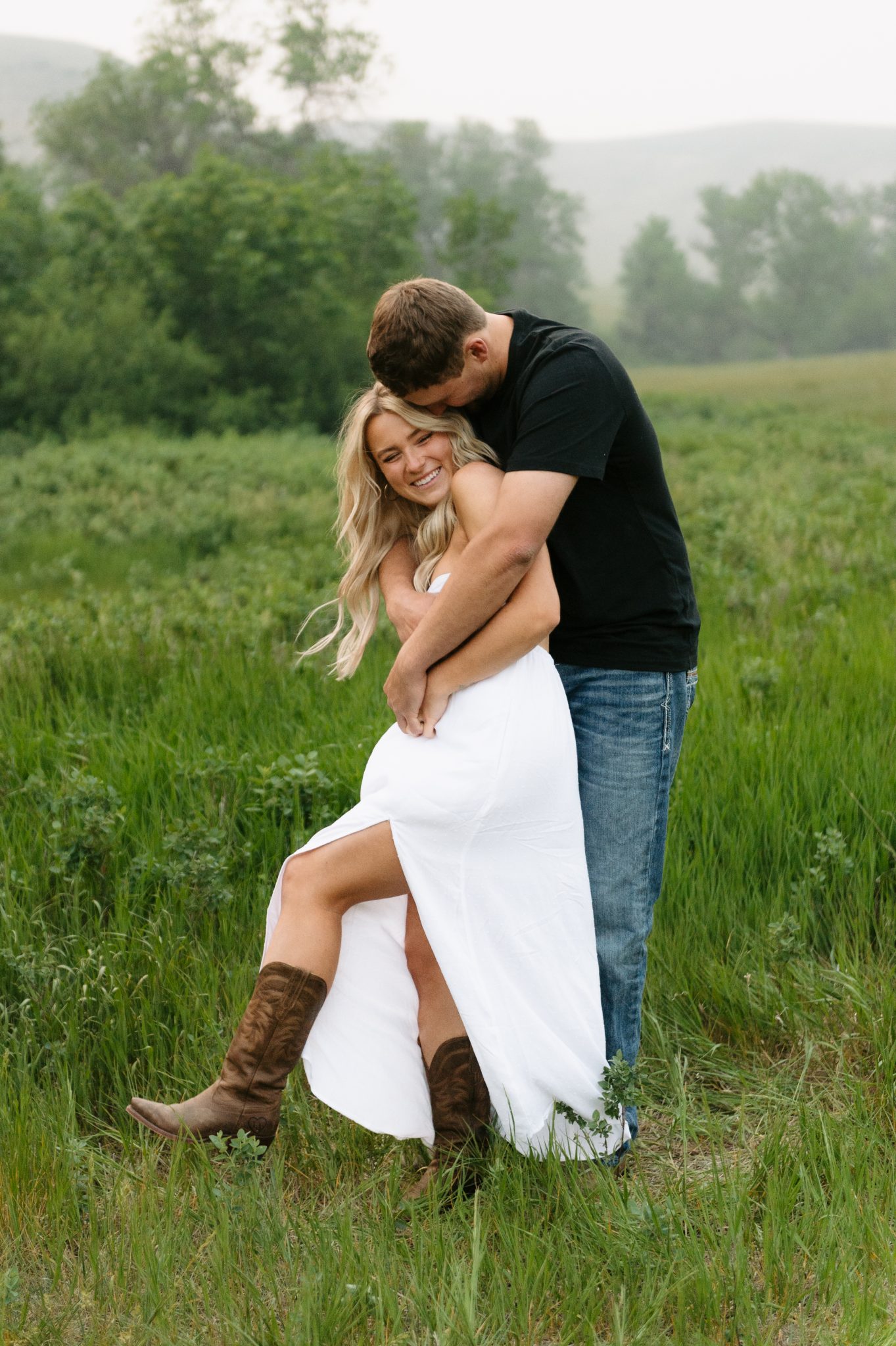 Couple laughing and embracing in a lush green meadow during a Scobey Montana engagement session rocky mountain bride.