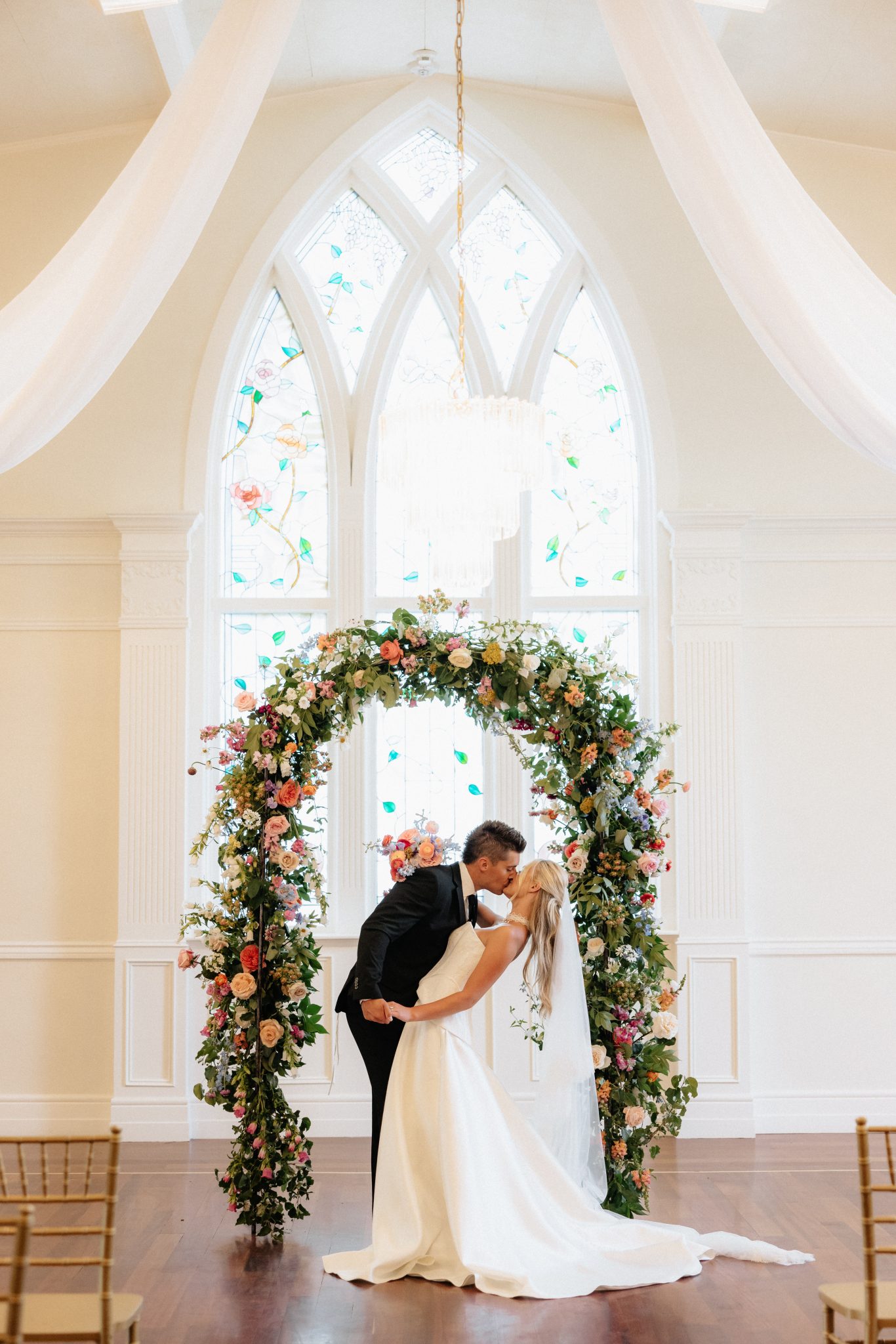 Bride and groom sharing a kiss under a lush floral arch with stained glass windows in the background, captured for The Northampton House and Jenny Pearl Photography wedding inspiration rocky mountain bride.
