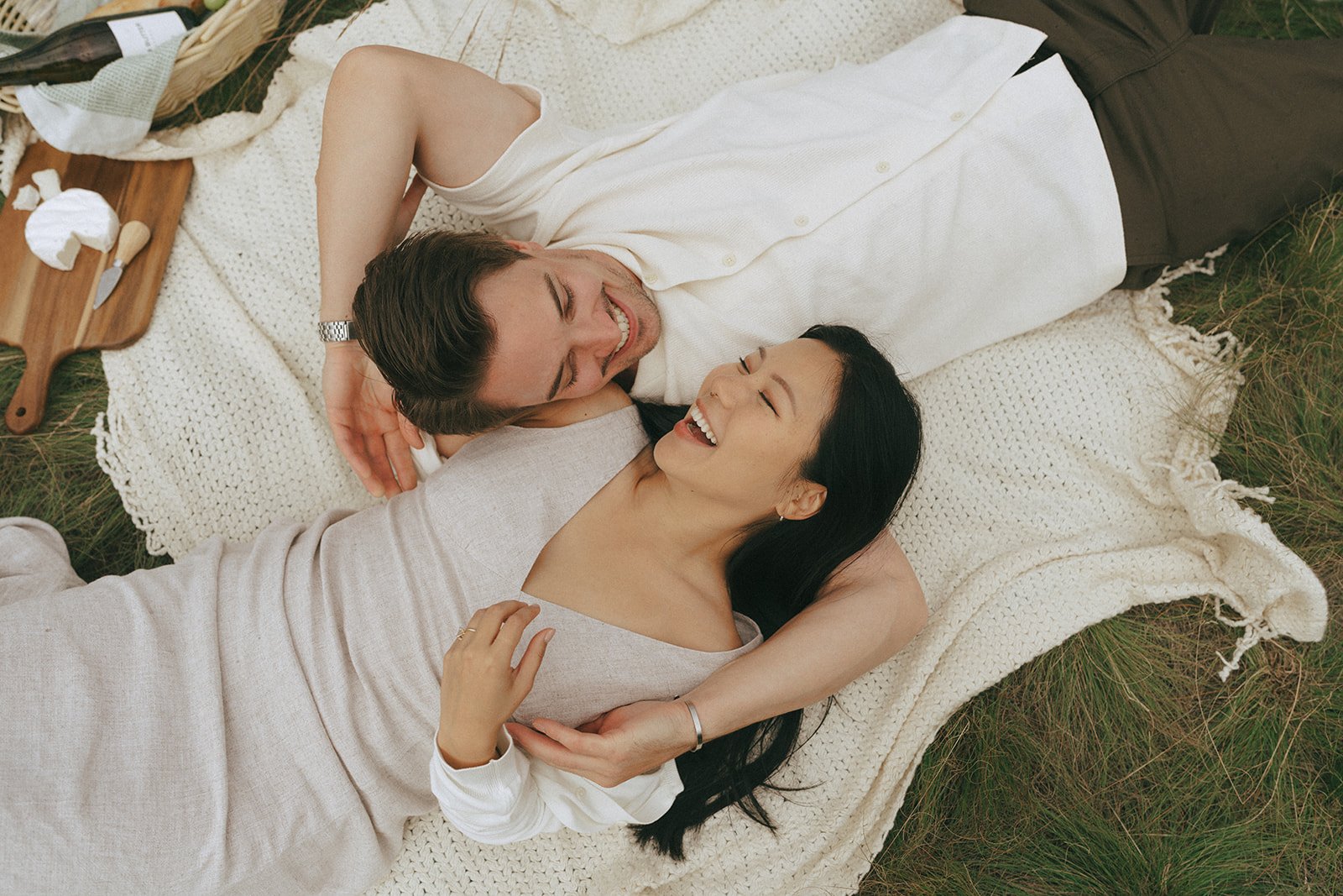 A couple lies on a cream blanket in the grass, laughing together during their canmore mountain quarry lake albert engagement taylor dawning photography session.