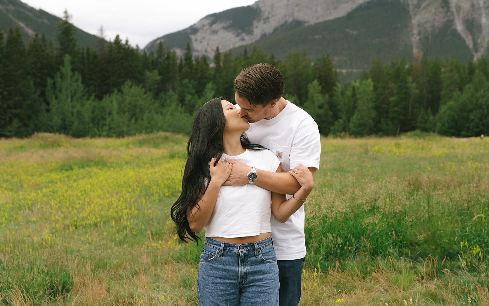 A couple shares a romantic kiss surrounded by tall pines and mountain peaks during their canmore mountain quarry lake albert engagement taylor dawning photography session.