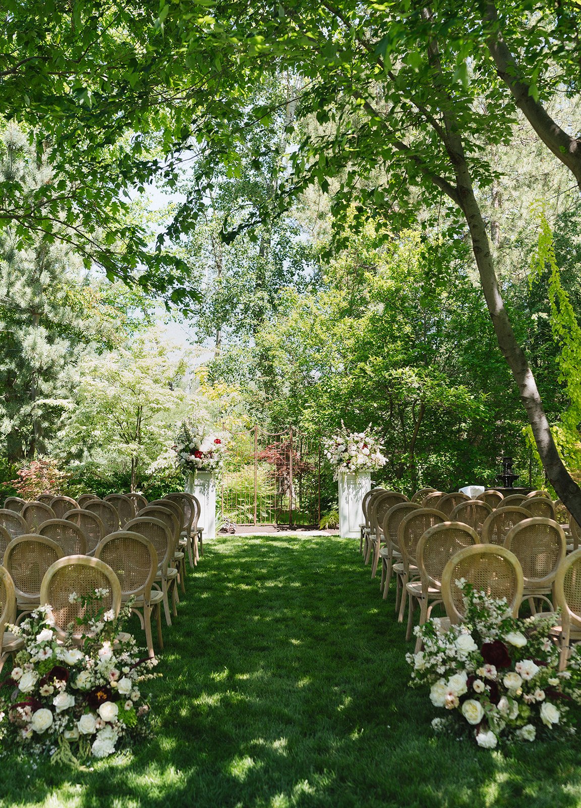 Ceremony aisle lined with chairs and lush floral arrangements under tall trees at a boise idaho private home intimate spring wedding emily battles photography.