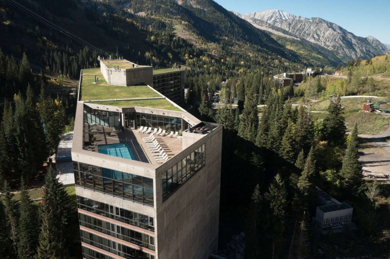 Aerial view of Cliff Lodge in summer with a rooftop pool, pine trees and dramatic mountain views at Snowbird in Utah.