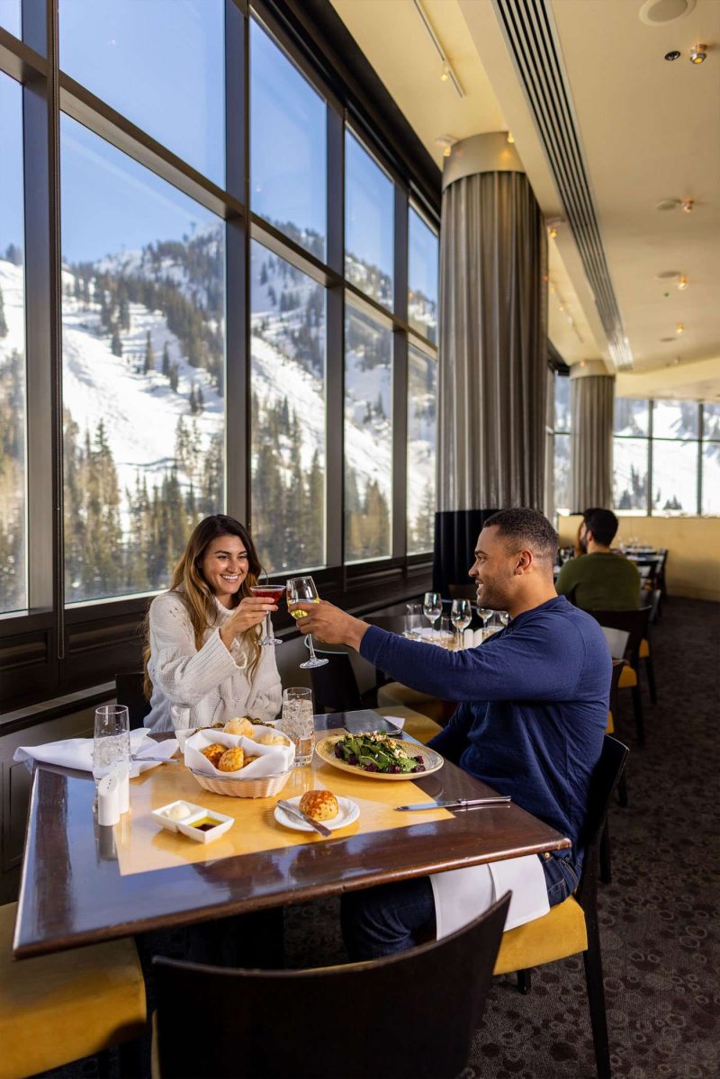 Couple toasting cocktails over lunch at The Aerie with floor-to-ceiling windows and snowy mountain views at Snowbird.