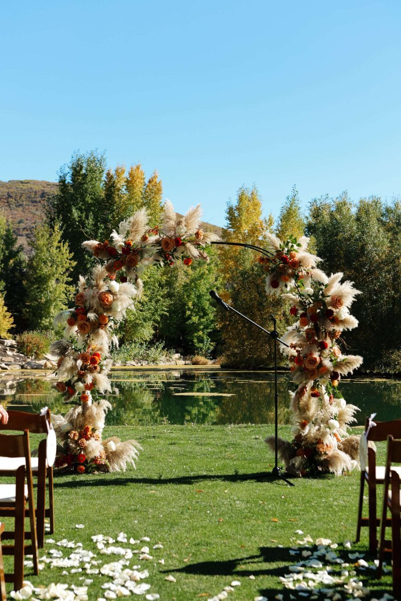 Outdoor wedding ceremony arch with pampas grass and rust-colored florals overlooking a pond at High Star Ranch in Utah.