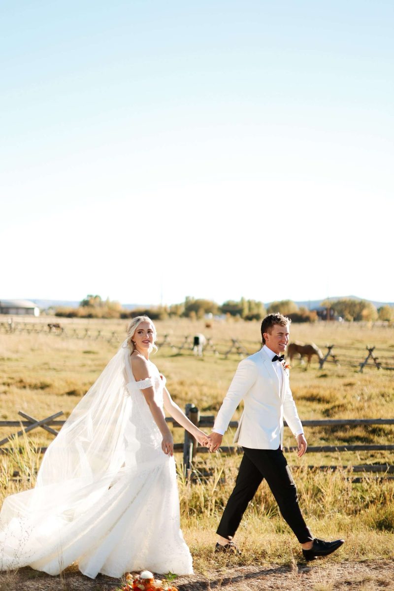 Bride and groom walking hand-in-hand through a golden ranch field with horses in the background at High Star Ranch.