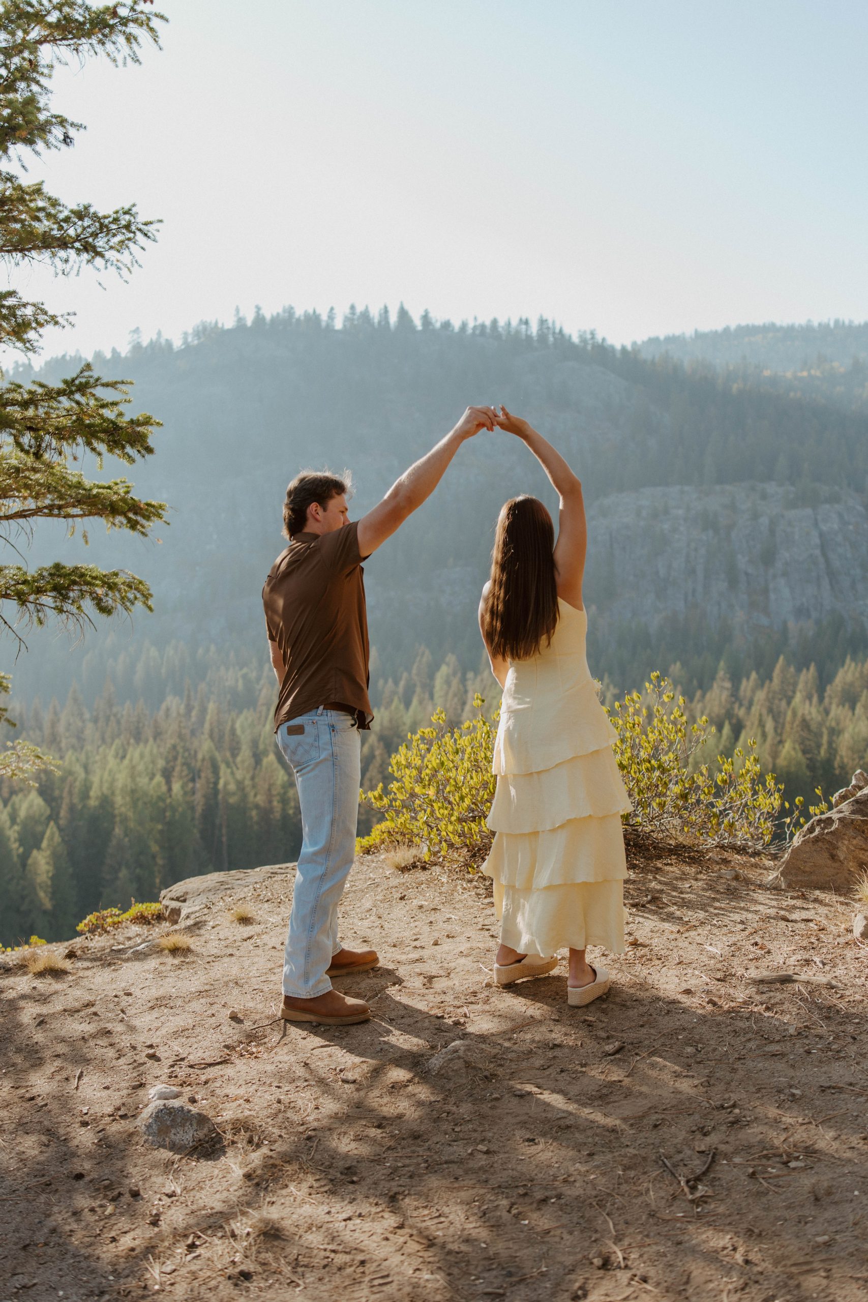 Couple holding hands and twirling on a cliff with panoramic forest views captured during a Quinn Briley Photography fall engagement session McCall Idaho rocky mountain bride.