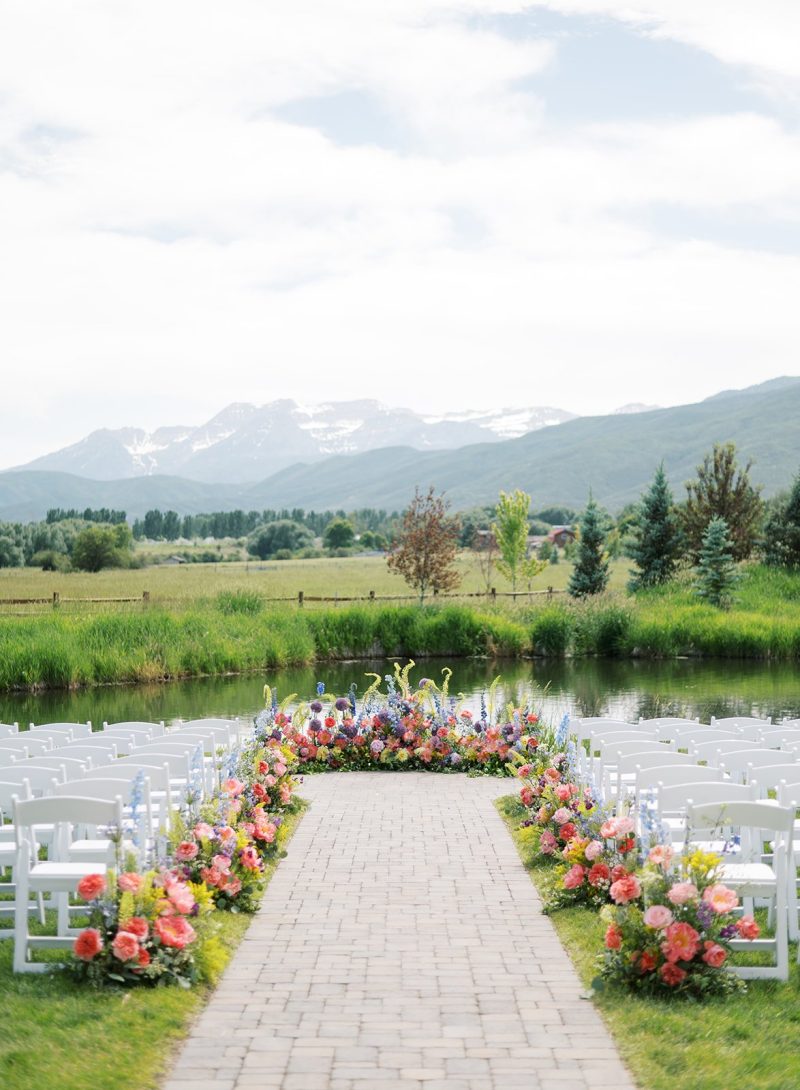 Colorful spring wedding ceremony aisle lined with bright florals, white chairs and pondside mountain views at River Bottoms Ranch.
