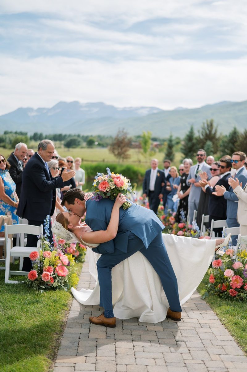 Bride and groom sharing a dip kiss during their River Bottoms Ranch wedding ceremony with guests cheering and colorful flowers lining the aisle.