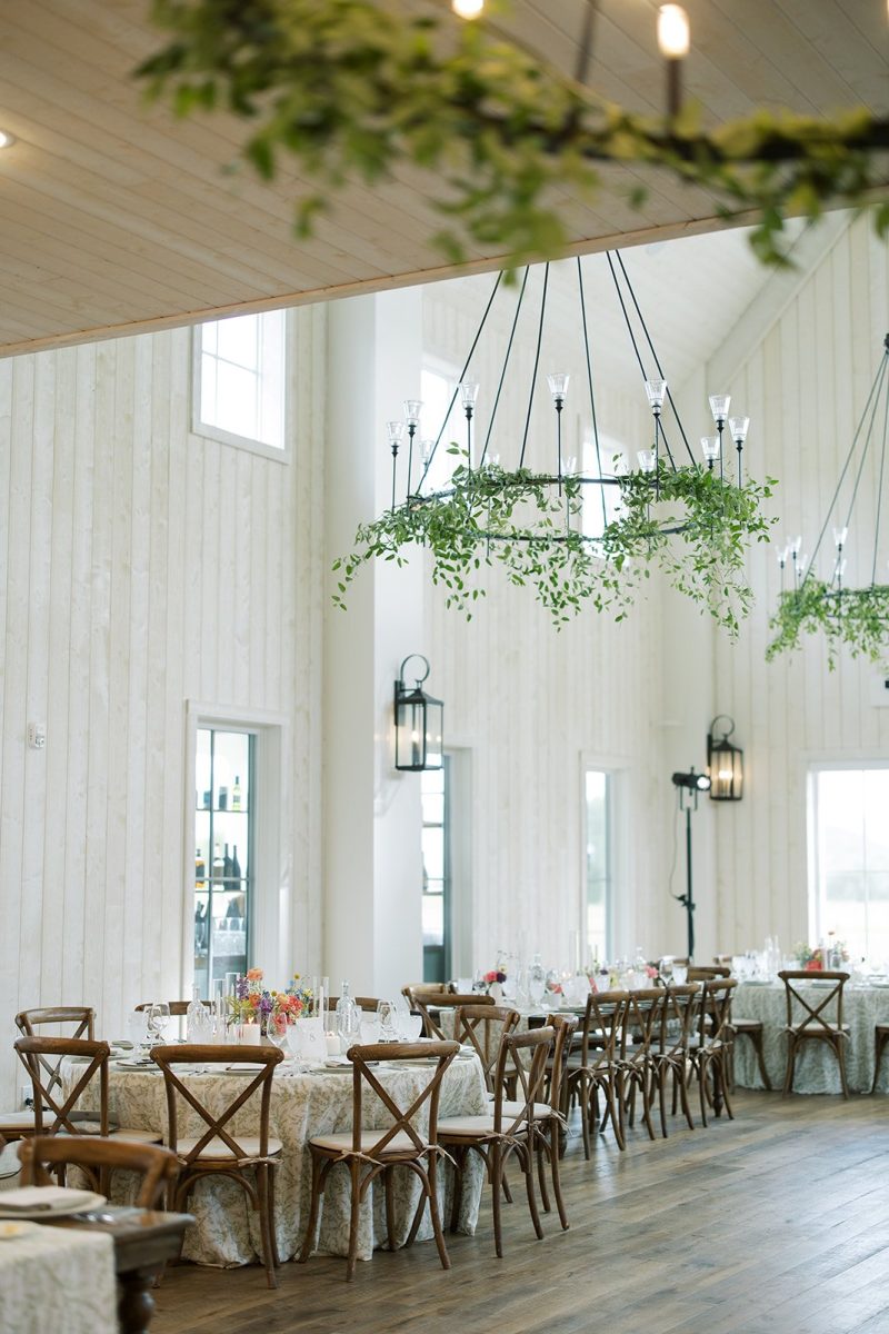 Bright indoor wedding reception at River Bottoms Ranch with greenery-wrapped chandeliers, wooden cross-back chairs and colorful floral centerpieces.