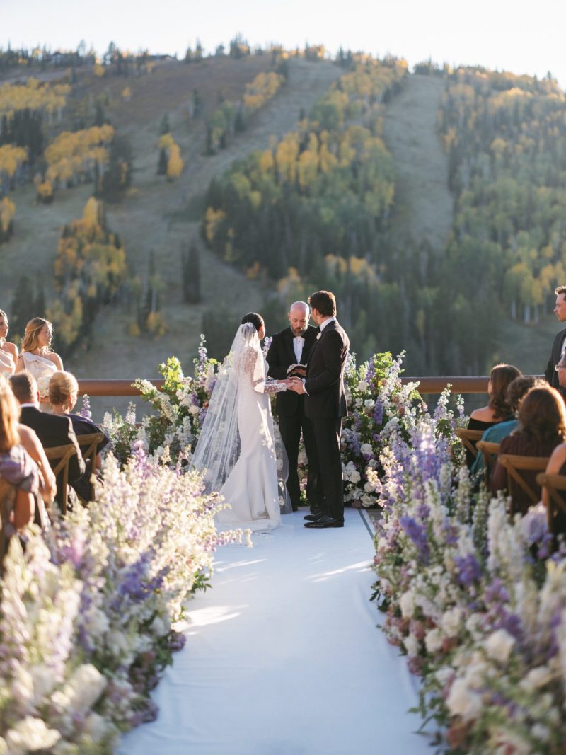Bride and groom exchanging vows during an outdoor Stein Eriksen Lodge wedding ceremony with a lush floral aisle and mountain views.