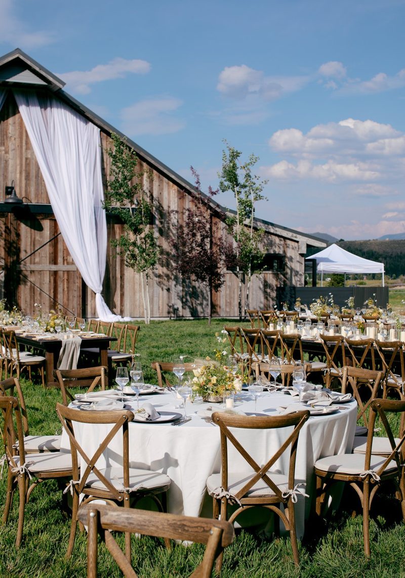 Outdoor summer wedding reception at 4U Ranch beside a rustic barn with white linens, wooden cross-back chairs, soft yellow florals and mountain views.