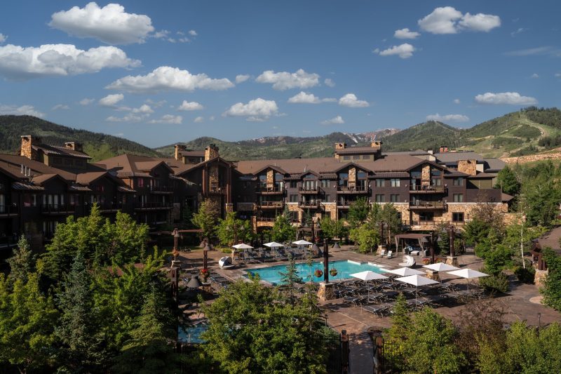 Aerial view of Waldorf Astoria Park City in summer with an outdoor pool, mountain lodge architecture and green Park City slopes in the background.