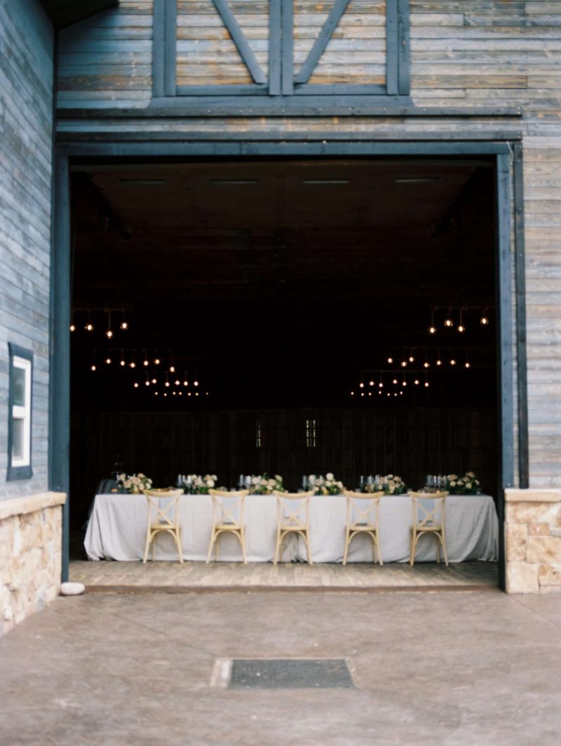 Rustic barn wedding reception with white linens, pale blush florals, cross-back chairs and warm hanging lights inside a Utah venue.