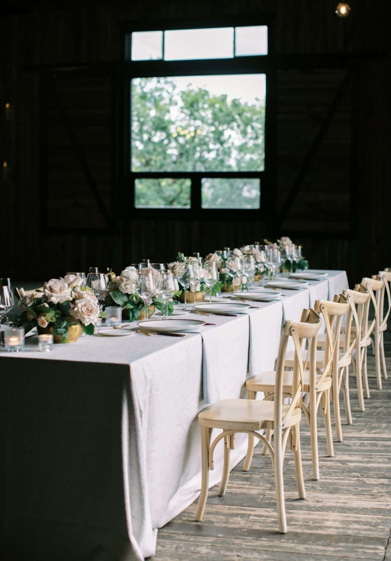 Long barn wedding reception table with neutral linens, candlelight, pale pink roses and light wood cross-back chairs.