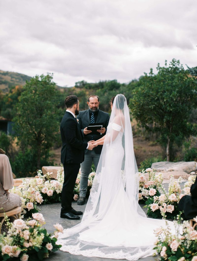Bride and groom exchanging vows during an outdoor Utah wedding ceremony surrounded by blush florals, greenery and mountain views.
